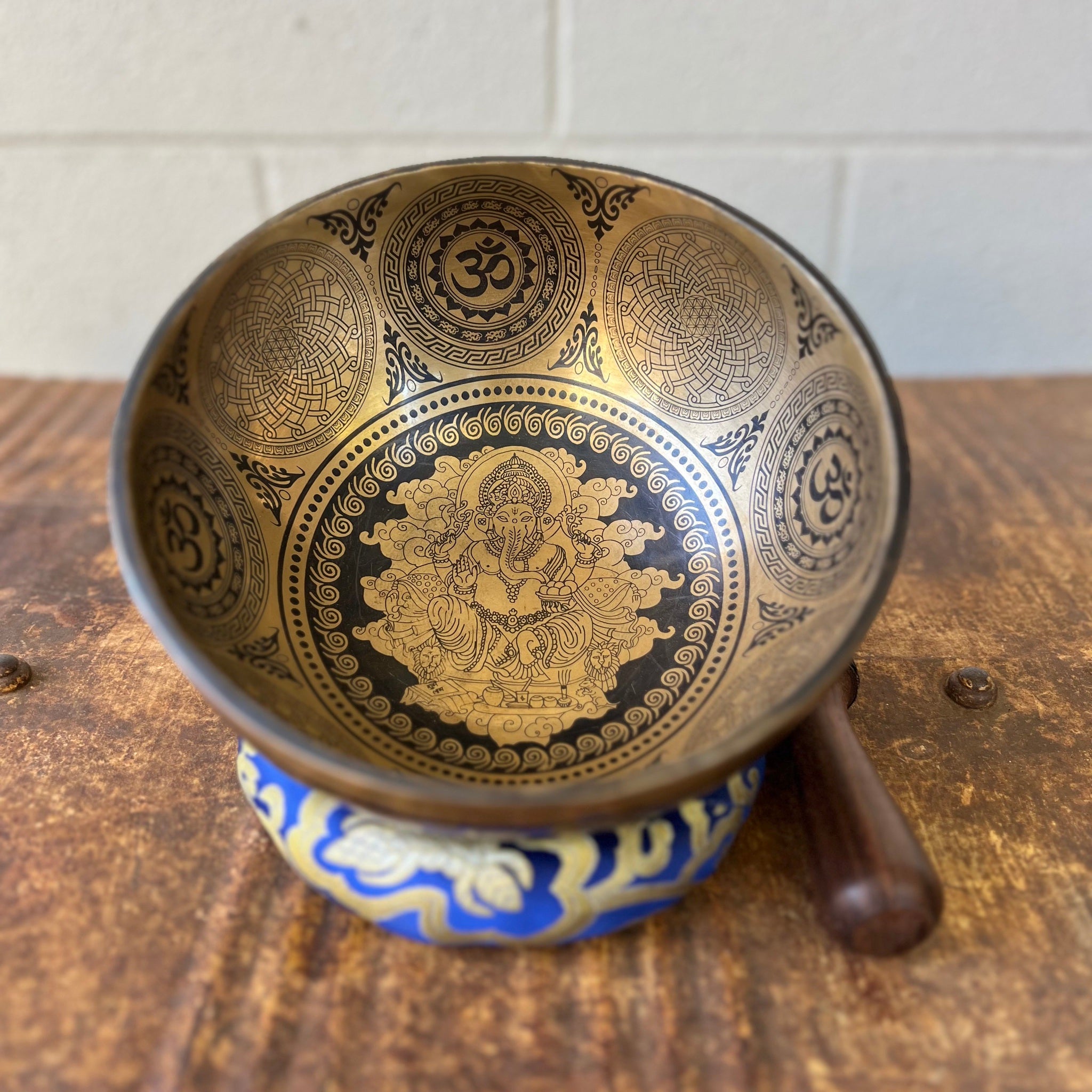 Bronze coloured 21cm singing bowl with black carving of Ganesh, Hindu OM symbol, and mandala designs, pictured with wooden striker and blue donut cushion