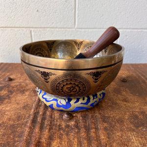 Bronze coloured 21cm singing bowl with black carving of Ganesh, Hindu OM symbol, and mandala designs, pictured with wooden striker and blue donut cushion