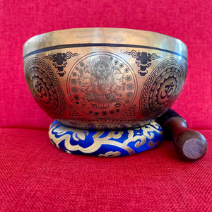 Bronze coloured 21cm singing bowl with black carving of Ganesh, Hindu OM symbol, and mandala designs, pictured with wooden striker and blue donut cushion