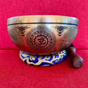 Bronze coloured 21cm singing bowl with black carving of Ganesh, Hindu OM symbol, and mandala designs, pictured with wooden striker and blue donut cushion