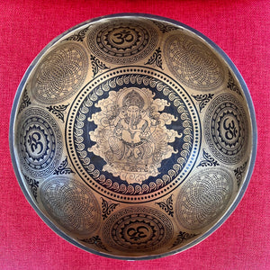 Bronze coloured 21cm singing bowl with black carving of Ganesh, Hindu OM symbol, and mandala designs, pictured with wooden striker and blue donut cushion
