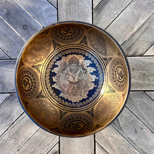 Bronze coloured 21cm singing bowl with black carving of Ganesh, Hindu OM symbol, and mandala designs, pictured with wooden striker and blue donut cushion