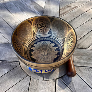 Bronze coloured 21cm singing bowl with black carving of Ganesh, Hindu OM symbol, and mandala designs, pictured with wooden striker and blue donut cushion